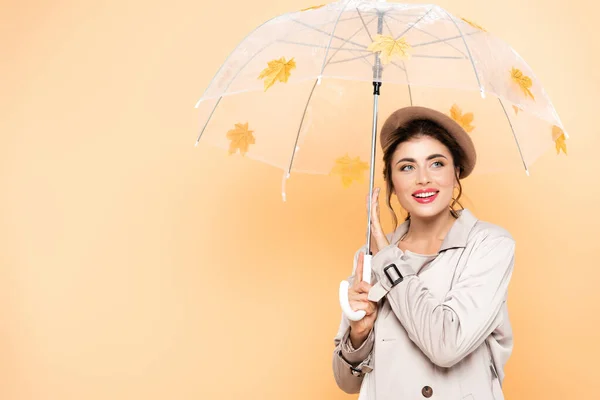 Fashionable woman in trench coat and beret posing under umbrella with yellow leaves on peach — Stock Photo