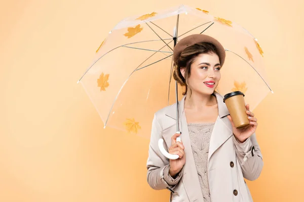 Trendy woman in beret and trench coat holding coffee to go under umbrella with autumn leaves on peach — Stock Photo