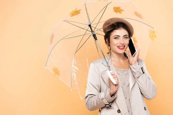 Joyful woman in fashionable autumn outfit talking on mobile phone under umbrella decorated with yellow leaves on peach — Stock Photo