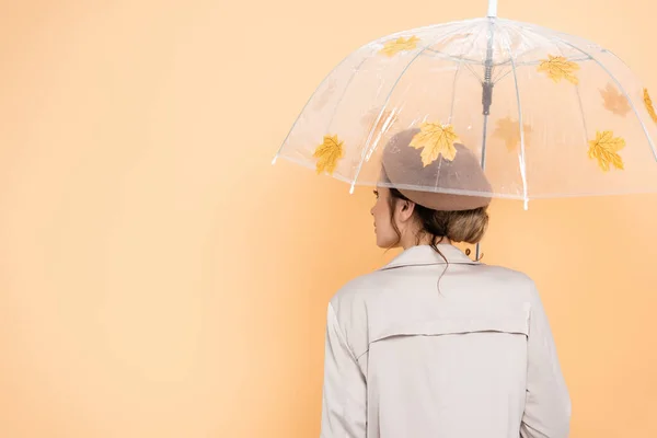 Back view of trendy woman in beret and trench coat under umbrella, decorated with yellow leaves on peach — Stock Photo
