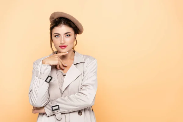 Fashionable woman in trench coat and beret touching chin while looking at camera on peach — Stock Photo