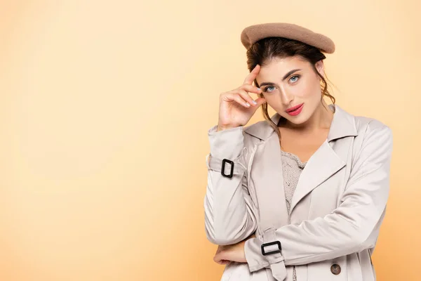 Femme à la mode en trench coat et béret touchant le front tout en regardant la caméra sur la pêche — Photo de stock