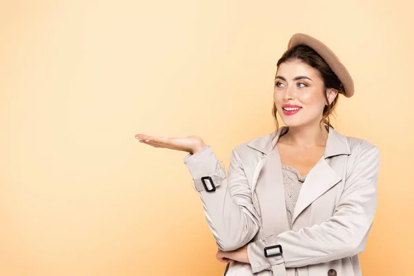 Fashionable woman in trench coat and beret pointing with hand on peach — Stock Photo