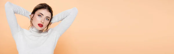Panoramic crop of woman in turtleneck holding hands behind head while posing on peach — Stock Photo