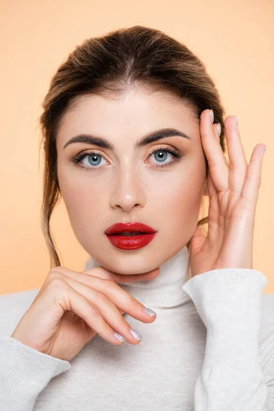 Mujer elegante tocando la cara mientras mira la cámara aislada en melocotón - foto de stock