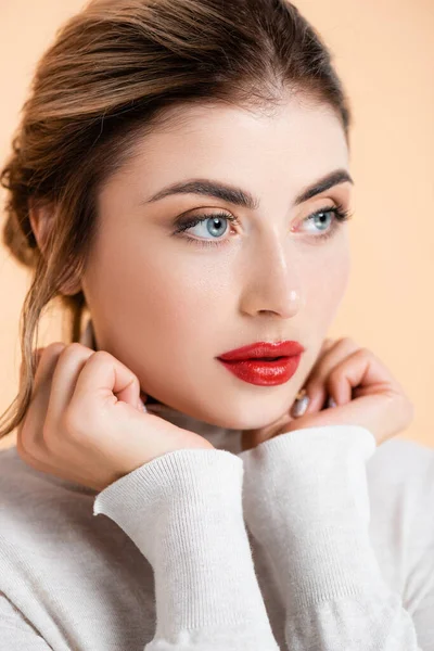 Mujer joven con labios rojos cogidos de la mano cerca de la cara mientras mira hacia otro lado aislado en melocotón - foto de stock