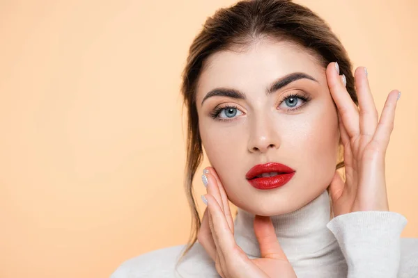 Sinnliche Frau mit roten Lippen, die in die Kamera schaut, während sie Gesicht isoliert auf Pfirsich berührt — Stockfoto