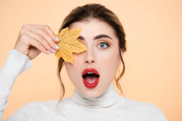 Excited woman with red lips looking at camera while covering eye with yellow leaf isolated on peach — Stock Photo