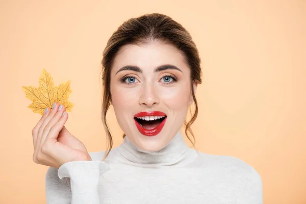 Mujer excitada y elegante con labios rojos sosteniendo la hoja amarilla mientras mira la cámara aislada en melocotón - foto de stock