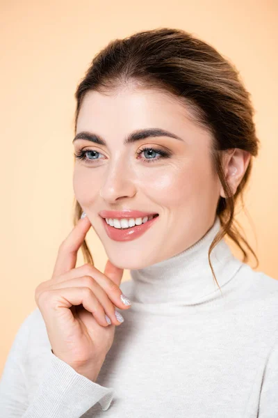 Freudige Frau im Rollkragen berührt Gesicht, während sie isoliert auf Pfirsich in die Kamera schaut — Stockfoto