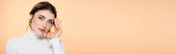 Horizontal image of young woman in turtleneck touching face while looking at camera isolated on peach — Stock Photo