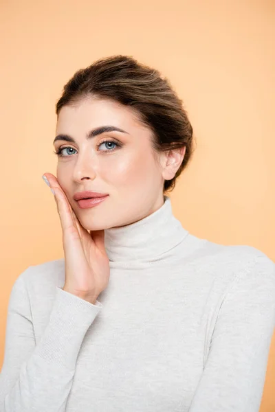 Sensual woman in turtleneck touching face while looking at camera isolated on peach — Stock Photo