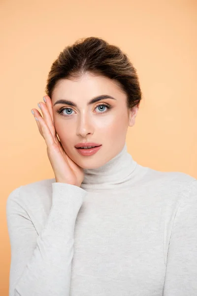 Young woman in turtleneck touching face while posing isolated on peach — Stock Photo