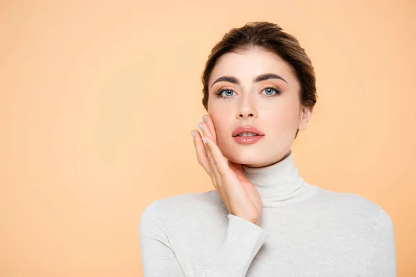 Woman in turtleneck looking at camera while touching face isolated on peach — Stock Photo