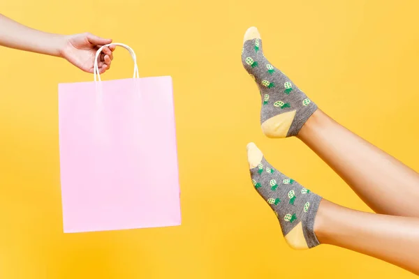 Vista recortada de las piernas femeninas en calcetines con impresión de piñas cerca de una bolsa de papel rosa aislada en amarillo - foto de stock