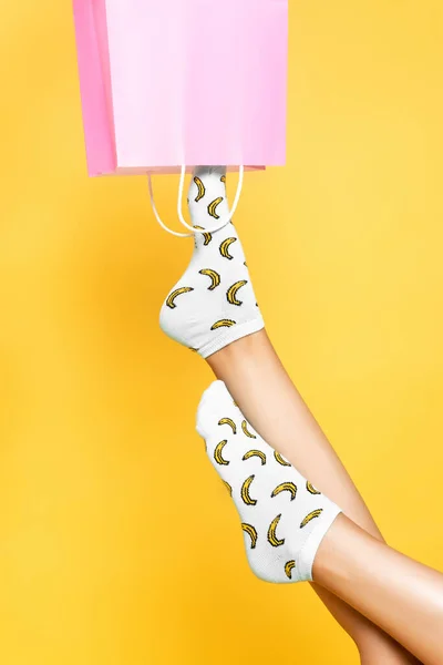 Vue recadrée de la femme portant des chaussettes, mettant la jambe dans un sac en papier rose isolé sur fond jaune — Photo de stock