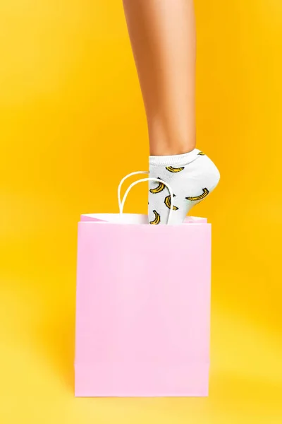 Vista recortada de mujer con calcetín poniendo pie en bolsa de papel rosa aislado sobre fondo amarillo - foto de stock