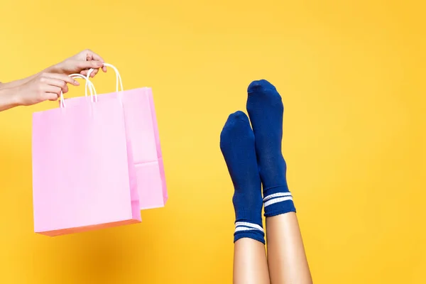 Vista recortada de la mujer sosteniendo bolsas de papel rosa cerca de las piernas femeninas en calcetines azules aislados sobre fondo amarillo - foto de stock