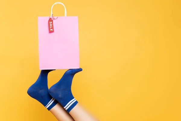 Visão superior das pernas femininas em meias azuis segurando saco de papel com etiqueta de venda isolada no fundo amarelo — Fotografia de Stock