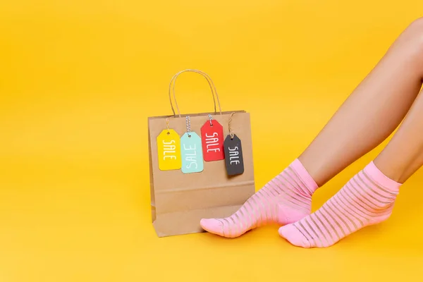 Обрізаний вид жіночих ніг в шкарпетках з рожевими смугами біля паперового мішка з мітками з продажу слова ізольовані на жовтому — стокове фото