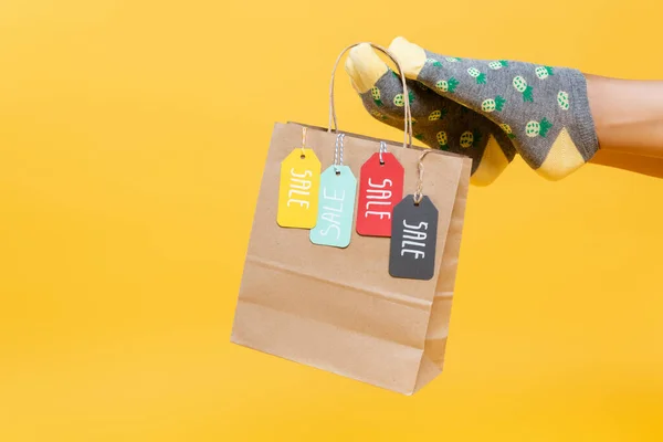 Cropped view of paper bag with sale tags hanging on woman leg in socks isolated on yellow — Stock Photo