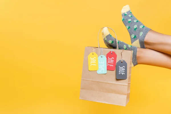 Cropped view of paper bag with sale tags hanging on woman legs in socks isolated on yellow — Stock Photo