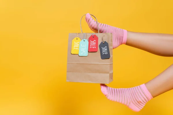 Vista recortada de la bolsa de papel con etiquetas de venta colgando de piernas de mujer en calcetines aislados en amarillo - foto de stock