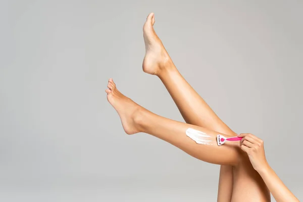 Обрезанный вид женщины с ногами в воздухе, бритья ноги с безопасной бритвой и кремом изолированы на сером фоне — стоковое фото