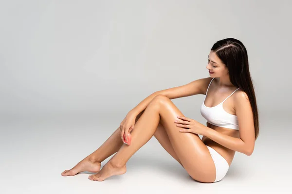 Brune femme en sous-vêtements blancs, jambe à raser avec épilateur rose, assise sur gris — Photo de stock