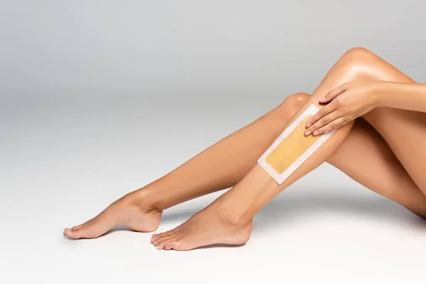 Cropped view of woman applying wax stripe on smooth leg on grey — Stock Photo