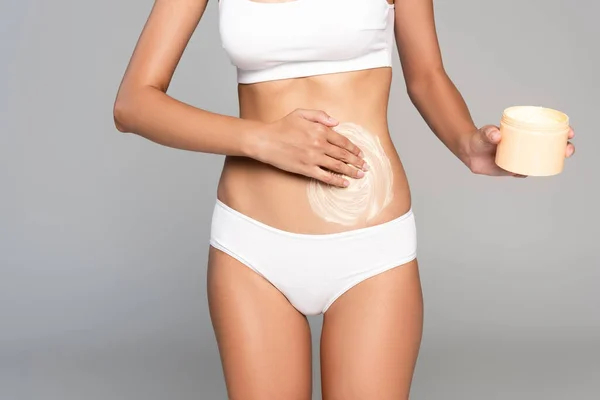 Cropped view of woman in white underwear holding jar and applying cream on belly isolated on grey — Stock Photo