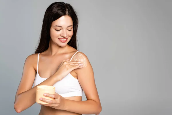 Mulher sorrindo aplicando creme no ombro e segurando frasco isolado em cinza — Fotografia de Stock