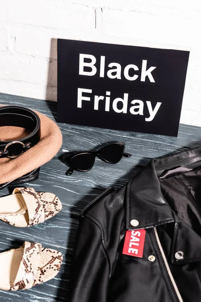 Zapatos, gafas de sol, boina y cinturón cerca de la tabla con el viernes negro y etiqueta con letras venta en la chaqueta de cuero - foto de stock