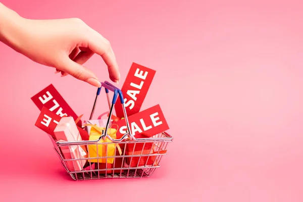 Cropped view of female hand near shopping basket with gifts and red tag with lettering on pink, black friday concept — Stock Photo