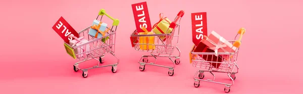 Panoramic crop of colorful presents in shopping carts with sale tags on pink, black friday concept — Stock Photo