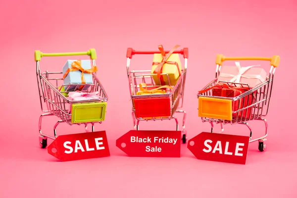 Colorful presents in shopping trolleys with sale tags on pink, black friday concept — Stock Photo