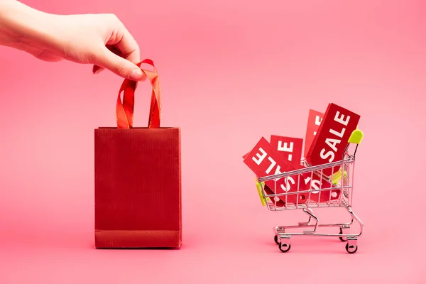 Cropped view of female hand touching shopping bag near cart with sale labels on pink — Stock Photo