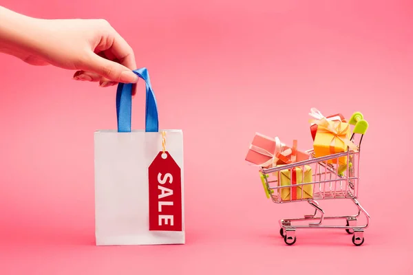 Ausgeschnittene Ansicht einer weiblichen Hand, die eine Einkaufstasche in der Nähe des Einkaufswagens mit Geschenken auf rosa hält — Stockfoto