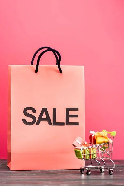 Shopping bag with sale lettering near small trolley with gifts on pink, black friday concept — Stock Photo
