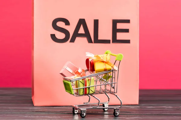 Selective focus of small trolley with gifts near paper bag with sale lettering on pink, black friday concept — Stock Photo