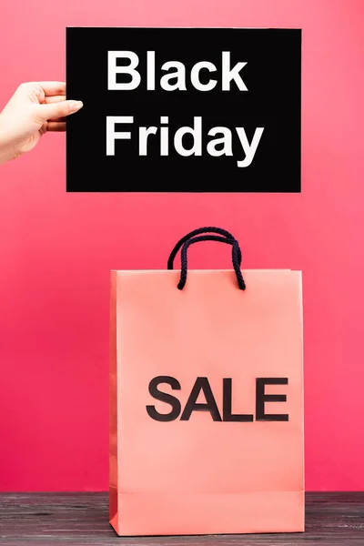 Ausgeschnittene Ansicht einer weiblichen Hand, die ein Plakat mit schwarzem Freitag-Schriftzug in der Nähe einer Einkaufstasche auf rosa hält — Stockfoto