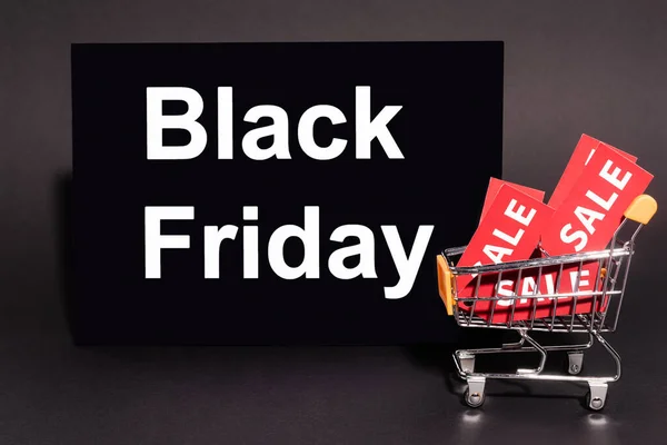 Placard with black friday lettering near toy cart with sale tags on dark background — Stock Photo