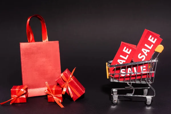 Shopping bag and gifts near toy cart with sale tags on dark background, black friday concept — Stock Photo