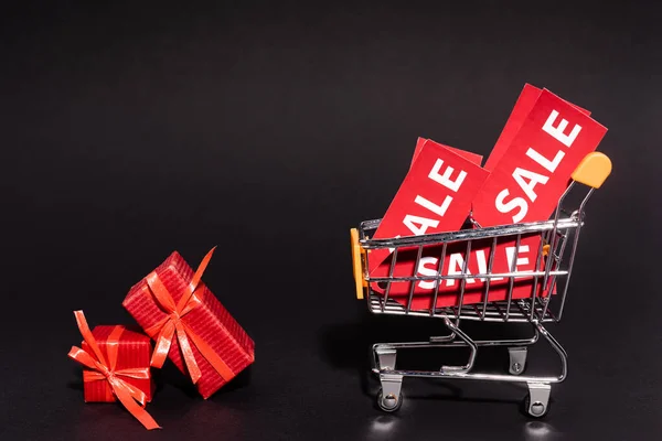 Cadeaux rouges près de chariot de jouets avec des étiquettes de vente sur fond sombre, concept vendredi noir — Photo de stock
