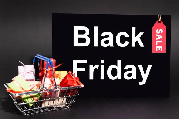 Small presents in toy shopping basket near placard with black friday lettering and sale tag on dark background — Stock Photo