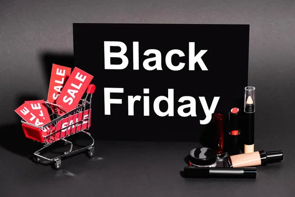 Placard with black friday lettering and toy cart with sale tags near decorative cosmetics on dark background — Stock Photo