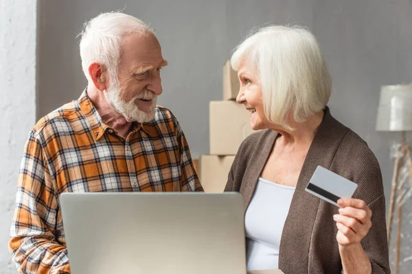 Sonriente pareja de ancianos haciendo la compra en línea utilizando el ordenador portátil y la tarjeta de crédito, mirándose entre sí - foto de stock