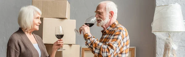 Panoramic shot of happy senior couple celebrating moving into new house with glasses of wine — Stock Photo