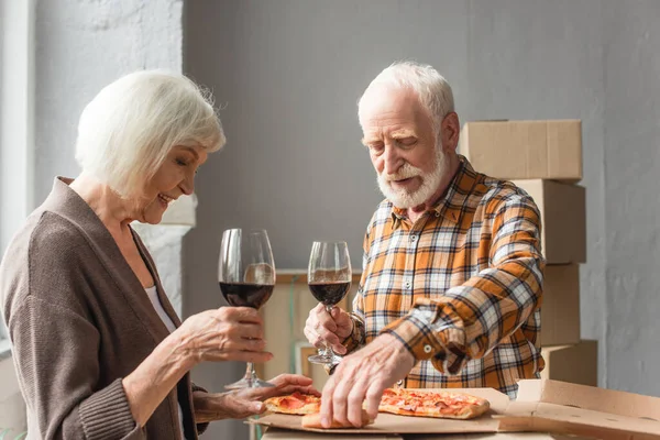 Coppia anziana che tiene bicchieri di vino e l'uomo che prende pezzo di pizza in una nuova casa — Foto stock