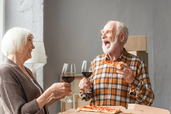 Lachender älterer Mann hält ein Stück Pizza in der Hand und schaut seine Frau an, während er ein Glas Wein in der Hand hält — Stockfoto
